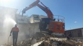 Large excavator demolishing a small building with urban demolition staff watching on 